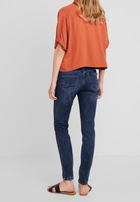 Camisa naranja corta con mangas enrolladas, combinada con jeans azul oscuro con una franja lateral. Sandalias planas completan el atuendo.