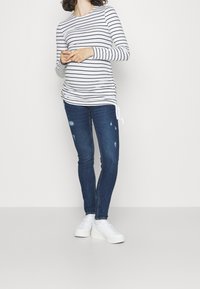 Randig långärmad topp i vitt och marinblått, med sidofåll, ihop med blå skinny jeans med slitna detaljer och vita sneakers.