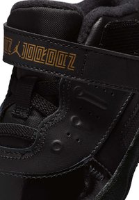 Zapatilla deportiva negra con puntera brillante, parte superior de malla texturizada, cuello acolchado y una correa decorativa dorada con la marca.