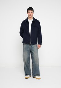 Tumši zila džinsa jaka ar rāvējslēdzēju un apkakli, priekšpusē kabata, valkāta pār baltu T-kreklu, kopā ar plaša piegriezuma izbalējušām džinsu biksēm un bēšām sporta kurpēm.