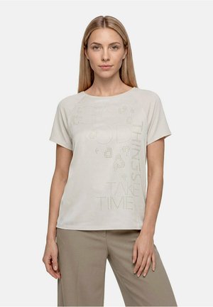 Femme blonde portant une chemise beige clair à manches courtes avec un texte perforé et un pantalon large taupe, debout devant un fond uni.