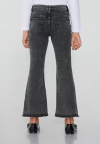 Černé džíny se zvýšeným pasem, rozšířenými nohavicemi a třepícími se lemy, se dvěma zadními kapsami. Hladká textura a rovný střih přes stehna.