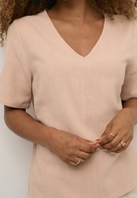 Blusa rosa chiaro con maniche corte, scollatura a V, tessuto strutturato e vestibilità ampia. Presenta dettagli di cucitura delicati lungo il centro.