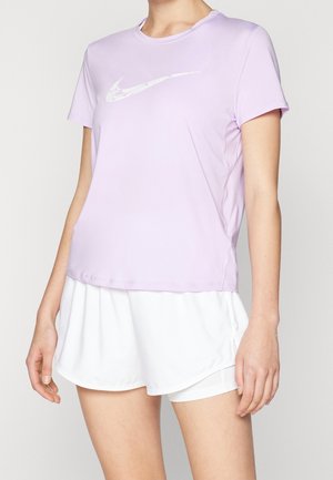 Personne portant un t-shirt Nike à manches courtes violet clair et un short de sport blanc sur un fond uni.
