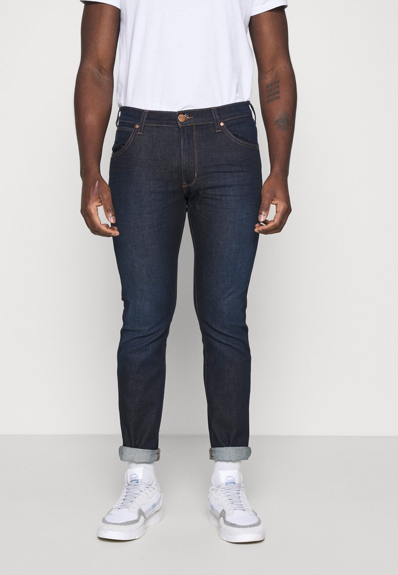 Jeans de denim oscuro con corte slim, con dobladillos enrollados, costuras naranjas en contraste y bolsillos clásicos. Combinados con una camiseta blanca y zapatillas.