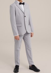 Ljusgrå kostym med en enkel knappstängning, snedskurna slag och sidofickor. Matchad med en svart fluga och svarta fin-skor.