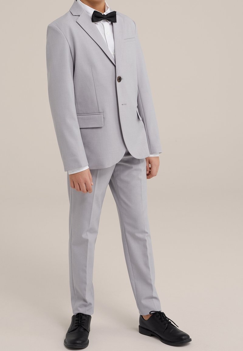 Ljusgrå kostym med en enkel knappstängning, snedskurna slag och sidofickor. Matchad med en svart fluga och svarta fin-skor.