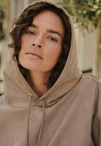 Taupe'owa bluza z kapturem o miękkiej teksturze, z troczkami i luźnym krojem, noszona przez modela na tle rozmytego krajobrazu na zewnątrz.