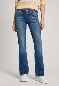 Medelblå jeans med vida ben, fem fickor, synlig sömnad och en något sliten yta. Kombinerat med vita sneakers.