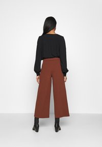 Černá halenka s dlouhým rukávem a nařaseným výstřihem spárovaná s hnědými širokými kalhotami s vysokým pasem. Modelka má na sobě černé kotníkové boty.