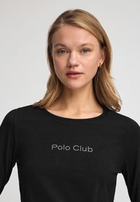 Schwarz langärmliges Shirt aus weichem Stoff mit dem Schriftzug "Polo Club" in metallischen Nieten auf der Brust. Klassisches Rundhalsdesign.