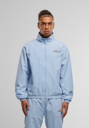 Helle blaue, leichte Jacke mit hohem Kragen, Reißverschluss, elastischen Bündchen und Branding auf der Brust. Das Material wirkt glatt und leicht glänzend.