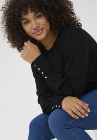 Black sweater with puffed sleeves and silver button details at the cuffs, worn with blue jeans. Background is plain white.