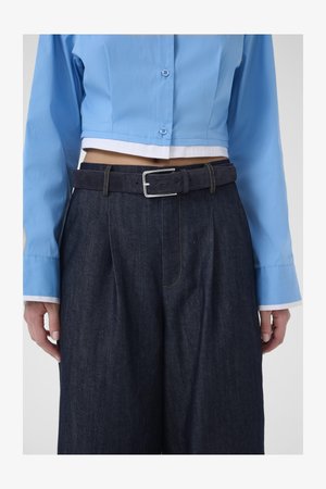 Īss zilas krāsas pogām aiztaisāms krekls, kopā ar tumši ziliem džinsa biksēm, kurās ir sudraba sprādzes josta un dzelteni izšūtas kabatas.