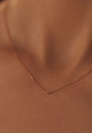 Collier constitué d'une délicate chaîne en or avec un design fin et enchaîné, reposant sur une surface de peau lisse et bronzée, mettant en valeur sa forme épurée et son éclat subtil.
