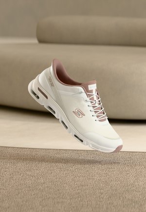 Baskets de sport blanches et mauves avec semelle à coussin d'air, logo Skechers sur le côté, exposées sur une surface texturée avec un canapé beige en arrière-plan.