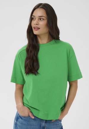 Femme aux longs cheveux foncés portant un t-shirt vert à manches courtes ample et un jean bleu, les mains partiellement dans les poches, regardant sur le côté.