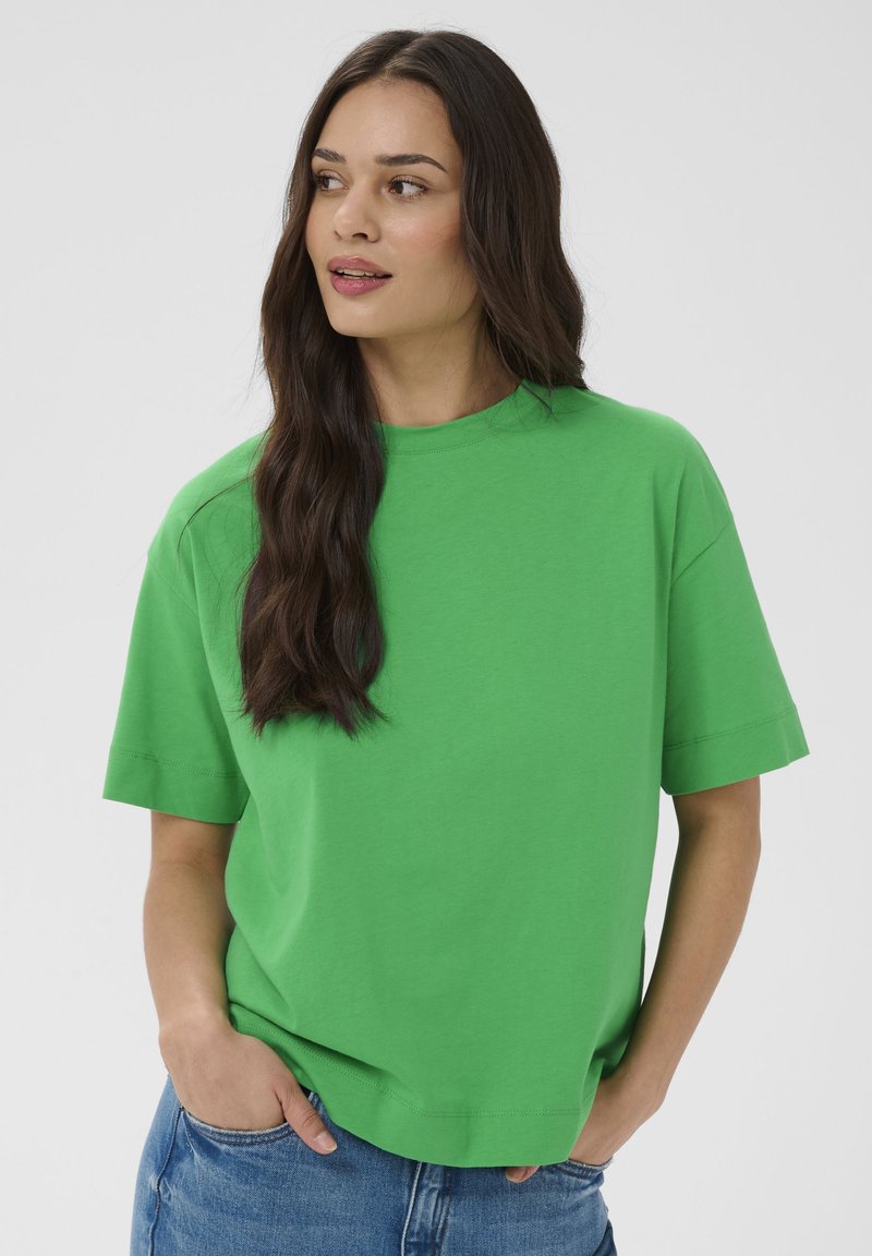Femme aux longs cheveux foncés portant un t-shirt vert à manches courtes ample et un jean bleu, les mains partiellement dans les poches, regardant sur le côté.