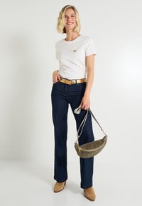 Camiseta blanca con un emblema de corazón dorado, jeans acampanados azul oscuro, botas de tacón beige y un bolso texturizado verde.
