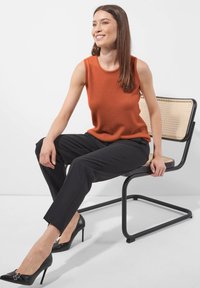 Eine Frau sitzt auf einem schwarzen Metall- und Rattanstuhl, trägt ein orangefarbenes ärmelloses Oberteil, schwarze Hosen und schwarze High Heels, lächelt und schaut nach rechts.