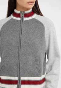 Cardigan grigio con zip e colletto alto a coste rosse, fascia in vita a righe bianche e rosse, e due tasche anteriori. Morbido tessuto a maglia.