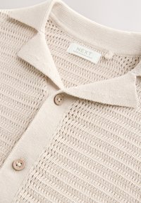 Beige strikket polo med tekstureret mønster, der har en blød krave og lukning med en enkelt knap. Labelen viser "NEXT, Made in Bangladesh."