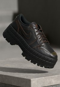 Ciemnobrązowy skórzany sportowy but Nike z grubą, masywną podeszwą z bieżnikiem, brązowymi przeszyciami i sznurówkami, unoszący się w powietrzu nad kamienną powierzchnią.