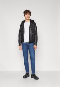 Chaqueta de cuero negra con cremalleras plateadas, superpuesta a una camisa blanca. Jeans ajustados azul y botas negras tipo tobillo completan el atuendo.