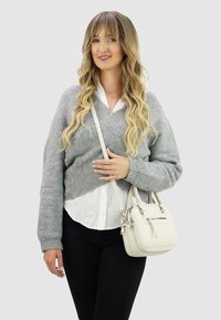 Jongere vrouw met lang golvend blond haar, gekleed in een grijze trui, witte blouse, zwarte broek, en houdt een witte handtas met een schouderband vast.