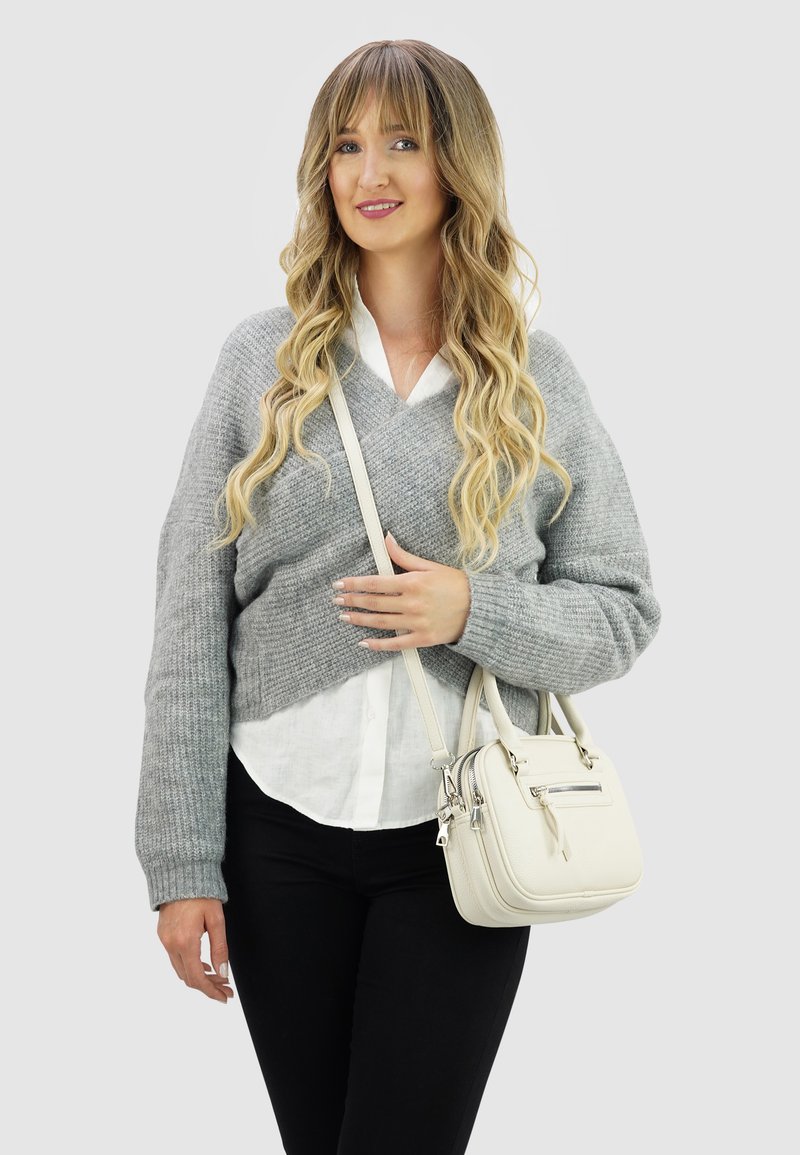 Jongere vrouw met lang golvend blond haar, gekleed in een grijze trui, witte blouse, zwarte broek, en houdt een witte handtas met een schouderband vast.