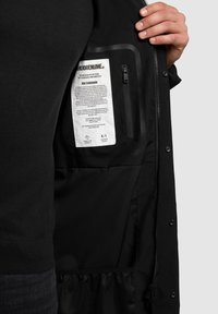 Zwarte buitenjas met een ritsvak aan de binnenkant en label. Heeft een gladde stoftextuur en een gaasvak aan de onderkant.