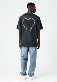 Melna pārskatāmā T-kreklā ar baltu tekstu, kas veido sirds formu uz aizmugures, valkāts ar gaiši ziliem džinsiem un baltām sporta kurpēm.