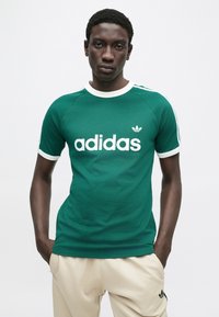Πράσινο t-shirt Adidas με λευκές λεπτομέρειες, με ένα εμφανές λευκό λογότυπο στο μπροστινό μέρος, κοντά μανίκια και στρογγυλή λαιμόκοψη.