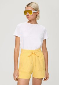 Witte katoenen t-shirt gecombineerd met gele badstof shorts met een elastische tailleband en een gestrikte voorkant, aangevuld met oversized gele zonnebril.