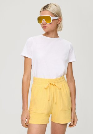 Weißes Baumwoll-T-Shirt kombiniert mit gelben Frottee-Shorts mit elastischem Bund und gebundenem Frontteil, ergänzt durch übergroße gelbe Sonnenbrillen.