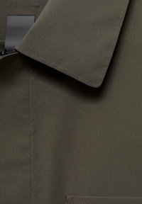 Tissu vert olive à texture lisse, avec un col plié et une poche plaquée, soulignés par des détails de couture subtils.