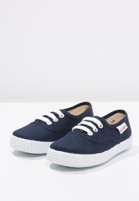 Zapatillas de lona navy con suela de goma blanca, que cuentan con cordones blancos, punta redonda y una pequeña etiqueta de logo en el lateral.
