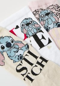 Három pár kötött zokni, amelyeken a Disney "Stitch" karaktere szerepel. A színek között fehér, rózsaszín és bézs található, különböző grafikai mintákkal és feliratokkal.