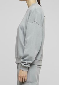 Sudadera gris claro de corte relajado, con puños y dobladillo acanalados, cuello redondo y detalles de costuras visibles en los hombros.