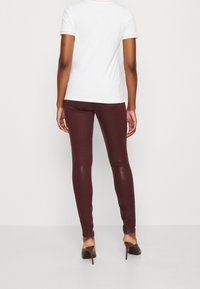 Kvinna står, vänd bort, iförd en vit kortärmad skjorta, vinröda tajta byxor och bruna högklackade skor.