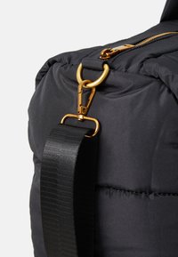 Black padded bag corner with gold-tone zipper, ring, and clasp attaching a wide black shoulder strap.