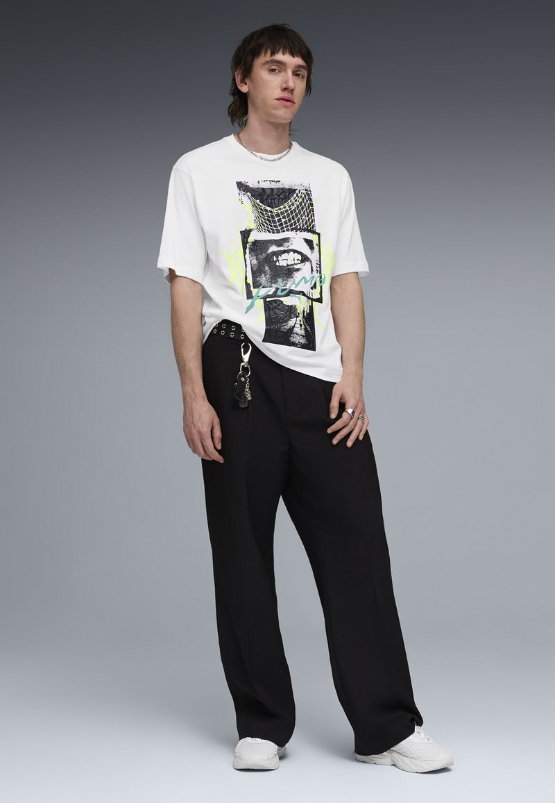 Jeune homme portant un t-shirt blanc à motifs, un pantalon large noir avec un porte-clés, des baskets blanches et des bagues argentées, sur un fond gris uni.