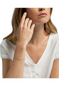 Guldkedja med flera dekorativa berlocker, som har en blandning av runda och ovala former, mot en ljus bakgrund och bärs på ett handled.