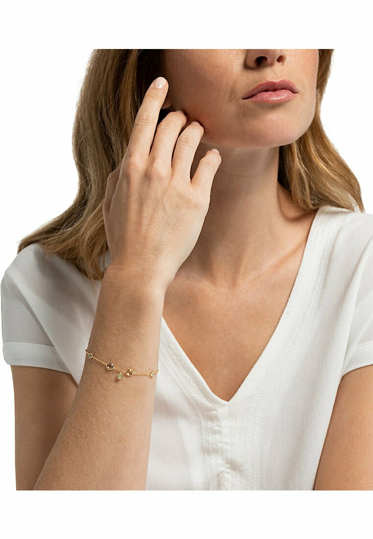 Guldkedja med flera dekorativa berlocker, som har en blandning av runda och ovala former, mot en ljus bakgrund och bärs på ett handled.