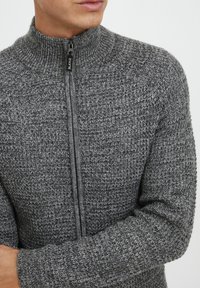 Grå strikket zip-up sweater med høj krave, der har et struktureret mønster og en mørk lynlås. Stoffet har et blødt og varmt udseende.