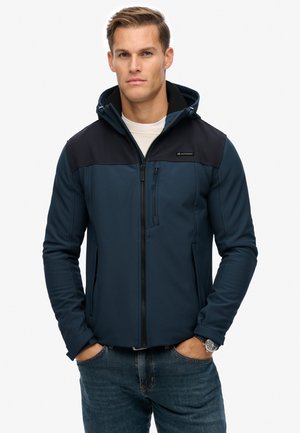 Marineblå softshell-jakke med et sort øverste panel, lynlåslukning, lynlåslommer og hætte. Glat tekstur, tætsiddende design.