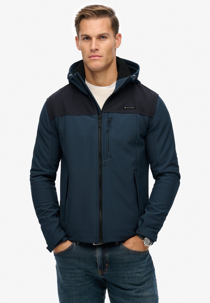 Dunkelblaues Softshell-Jackett mit einem schwarzen Oberteil, Reißverschluss, mit Reißverschluss versehenen Taschen und Kapuze. Glatte Textur, taillierte Passform.