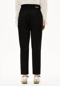 Jeans de mezclilla negra de tiro alto con pierna recta, con bolsillos traseros y una etiqueta en la cinturilla. Combinados con zapatillas blancas y verdes.