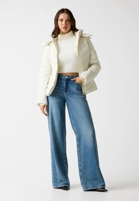 Giacca puffer bianca trapuntata con chiusura con zip e cappuccio, abbinata a jeans blu a vita alta e gamba larga, con tacchi a punta neri.