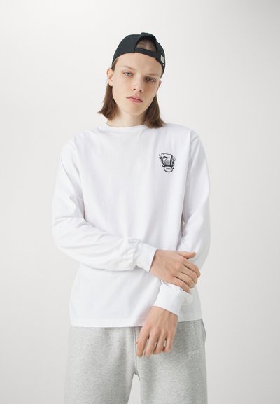 White long-sleeve shirt made of cotton, featuring a small black shield logo with the number 7. Worn with gray sweatpants, smooth texture.